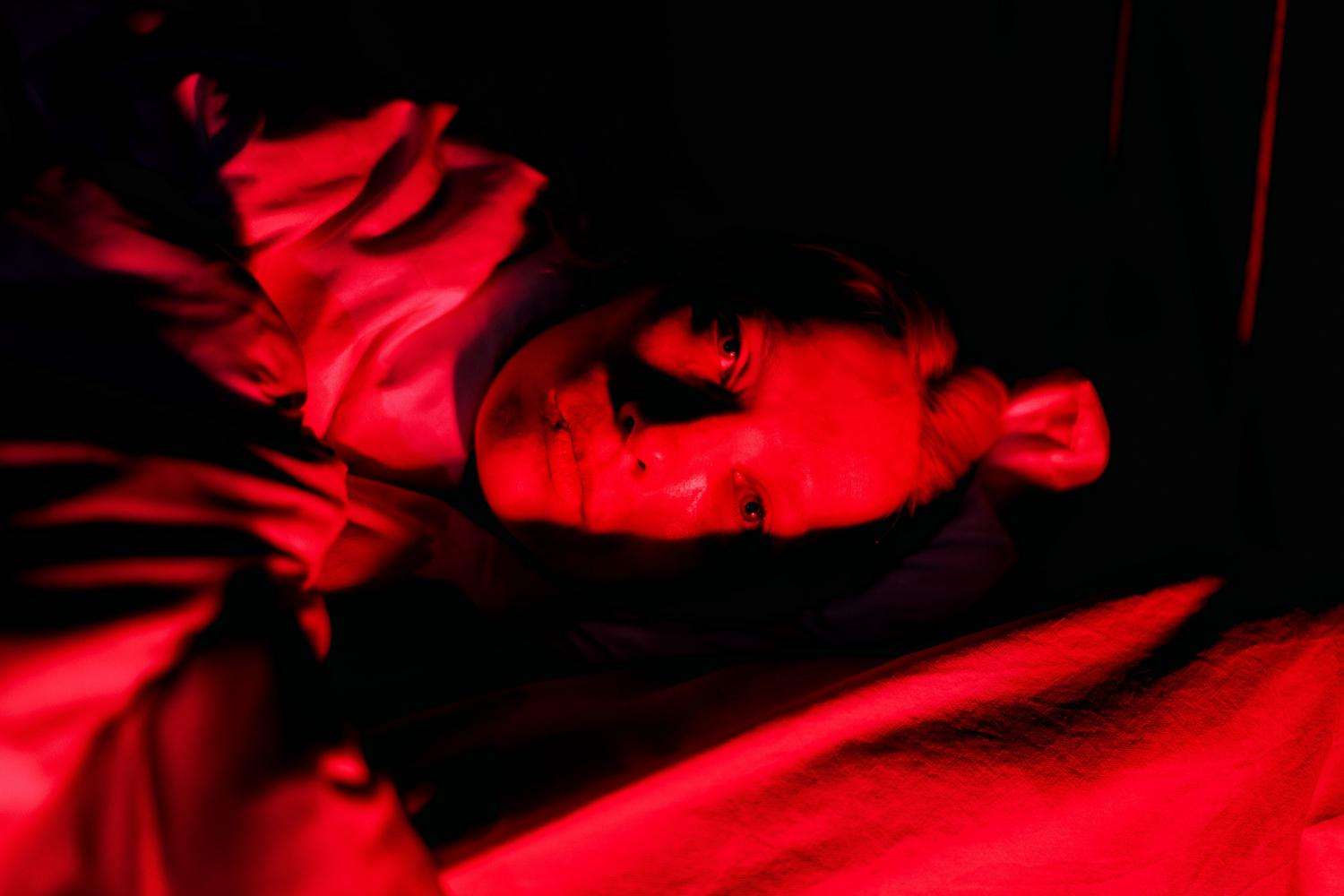 Retrato dramático con iluminación roja de una persona en la cama, simbolizando el estado de alerta permanente y el deterioro psicológico.